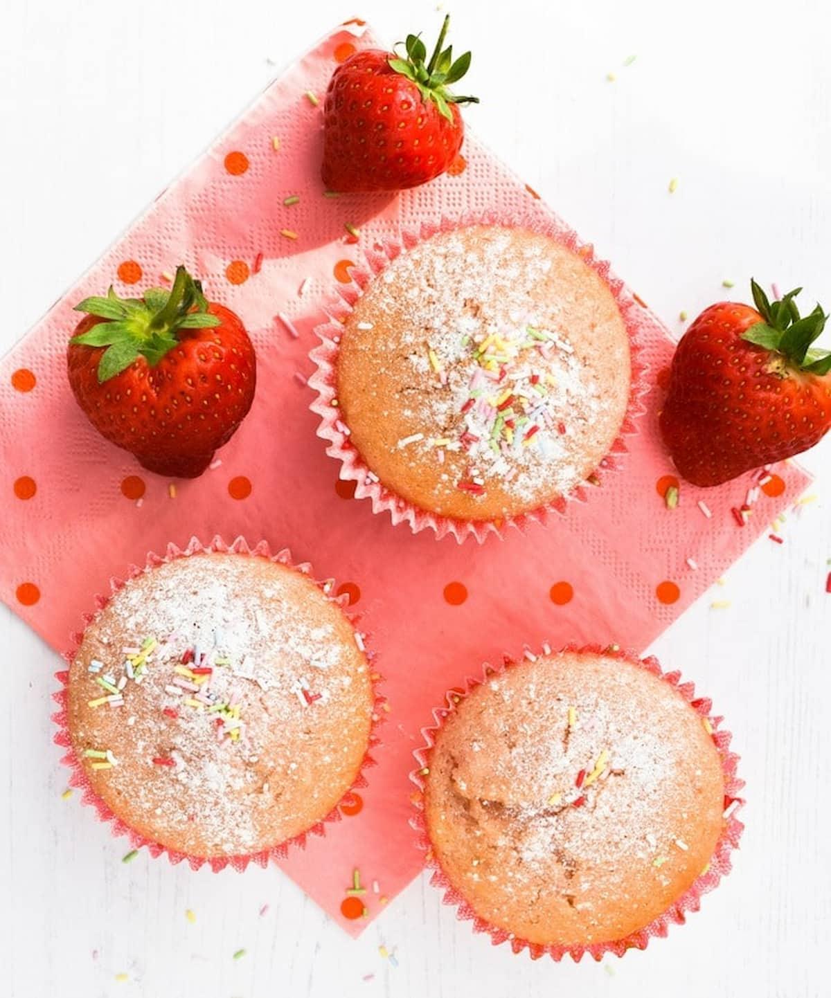 Strawberry muffins topped with sprinkles, next to fresh strawberries.