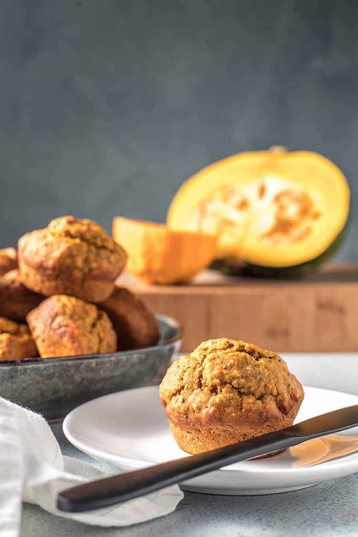Orange pumpkin muffin on a plate with more muffins a sliced pumpkin in the background.