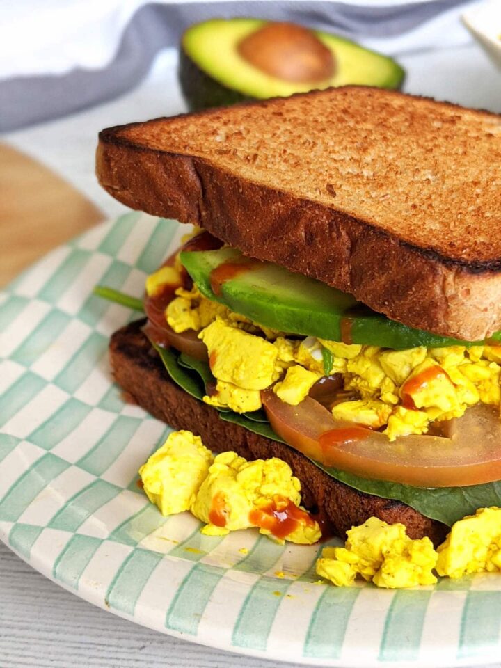 Silken tofu scramble served in a breakfast sandwich made with toast, spinach, tomatoes, sliced avocado, and a drizzle of hot sauce.