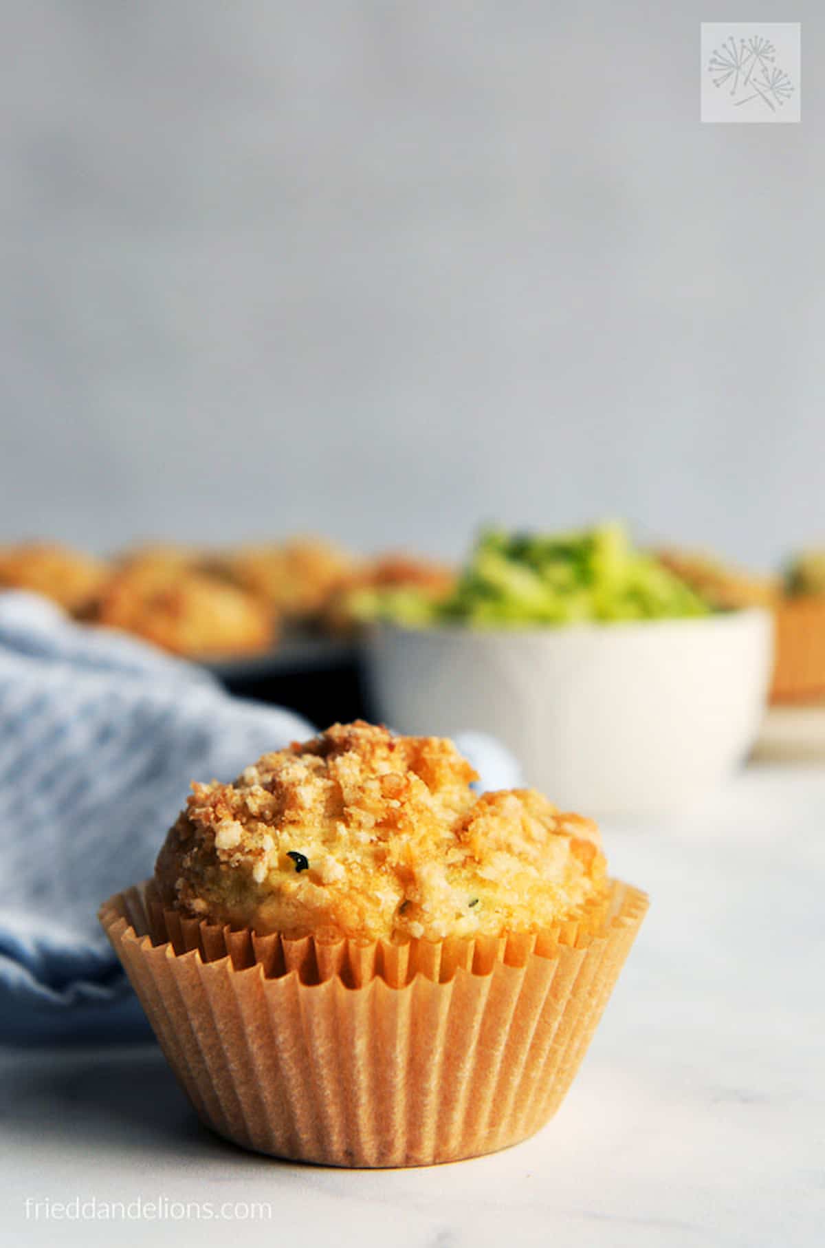 A coconut zucchini muffin in two muffin liners with a bowl of zucchini in the background.