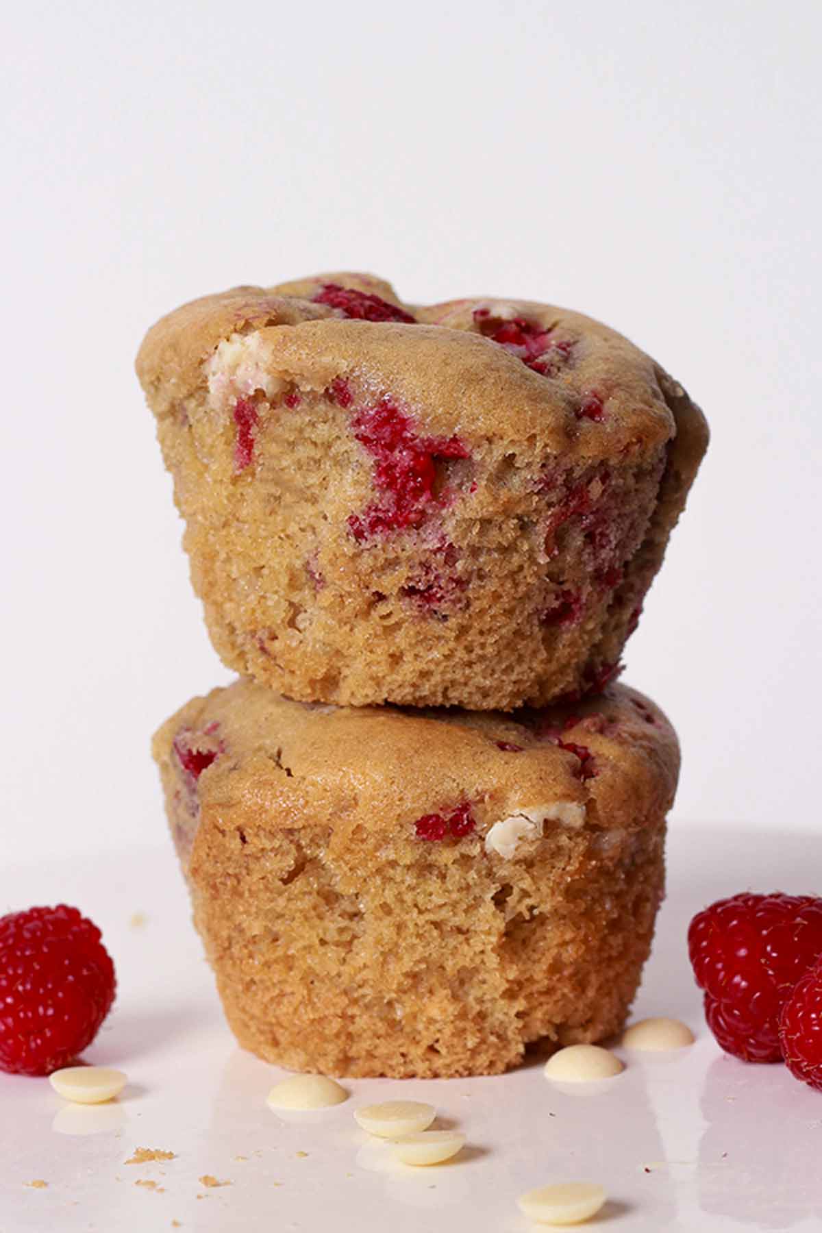 Two vegan raspberry white chocolate muffins stacked on one another, next to fresh raspberries.