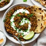 Chickpea and lentil curry served in a white bowl and topped with coconut yogurt and chopped cilantro. Two pieces of naan dipped on the side of the bowl.