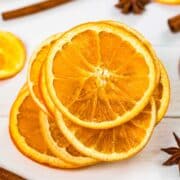A stack of dried orange slices on a white table next to star anise and cinnamon sticks.