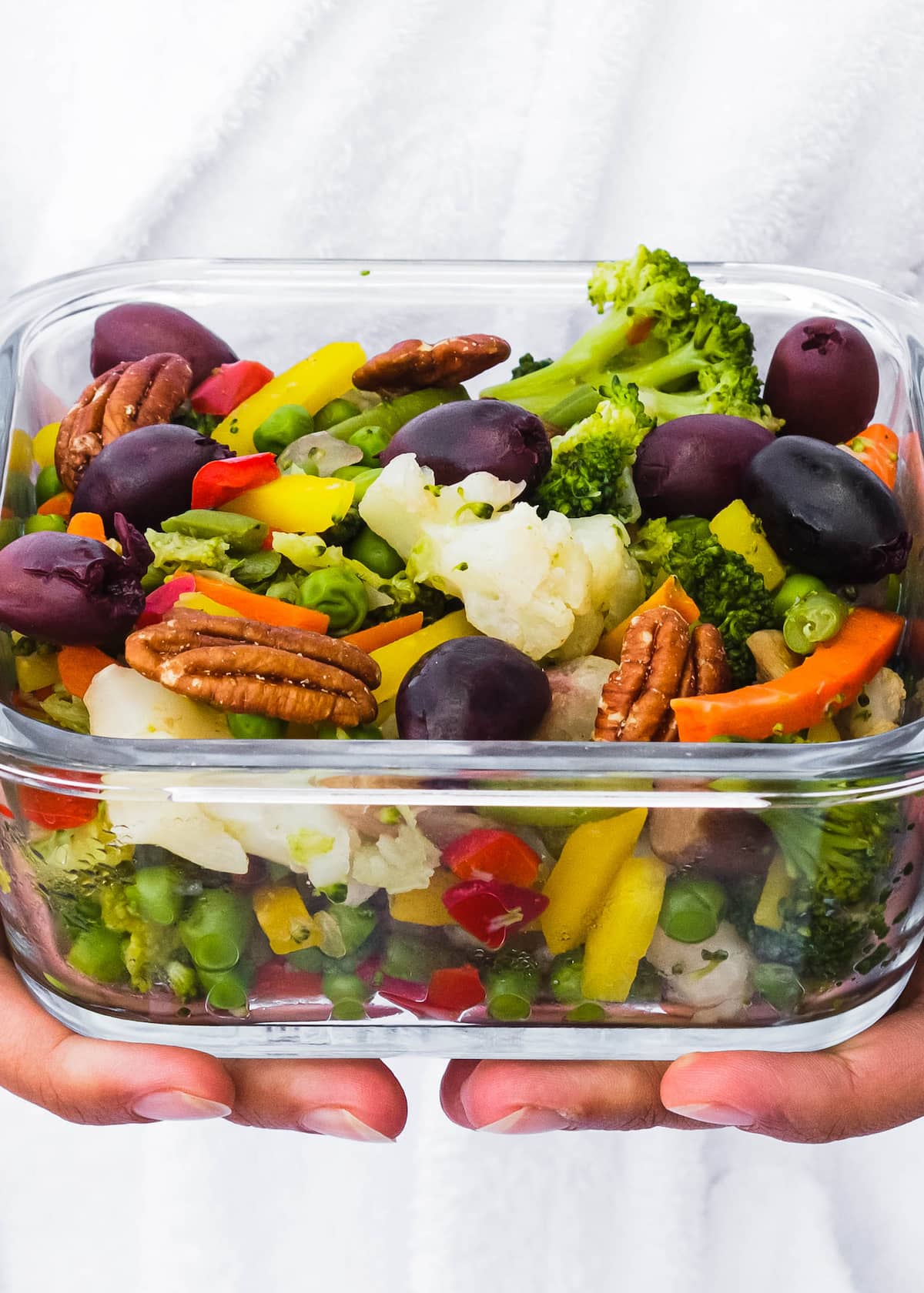 A clear glass container full of steamed veggies, pecans, and grapes.