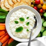 Vegan cottage cheese served in a white bowl and topped with fresh basil leaves. An array of vegetables surrounding the bowl