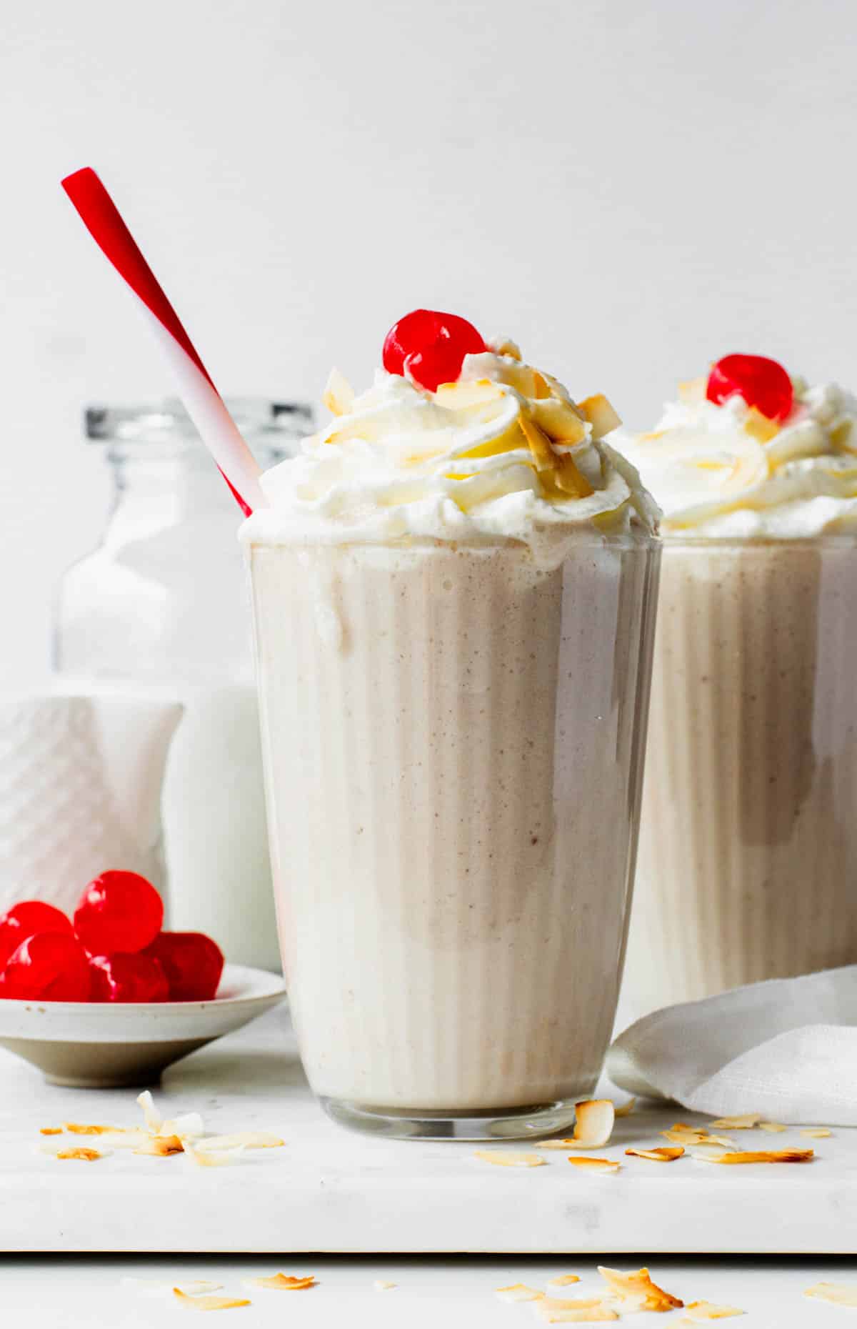 Two milkshake glasses filled with coconut shakes, topped with whipped cream and cherries.