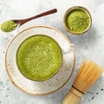 Whisked matcha in a bowl next to matcha powder, bamboo whisk, and teaspoon.