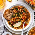Navy bean vegetable soup served in a white bowl with a slices of sourdough bread dipped in it.