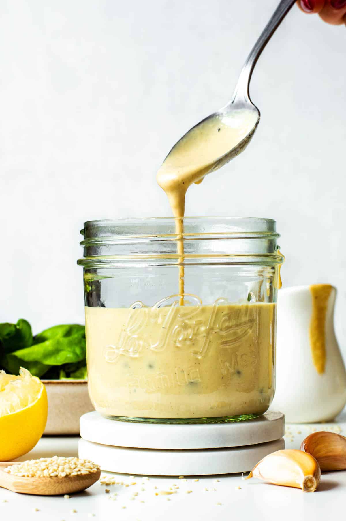 A spoon dripping tahini Caesar dressing into a glass jar.