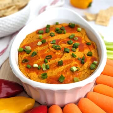 A small casserole dish of buffalo chickpea dip topped with green onion and served with fresh vegetables.