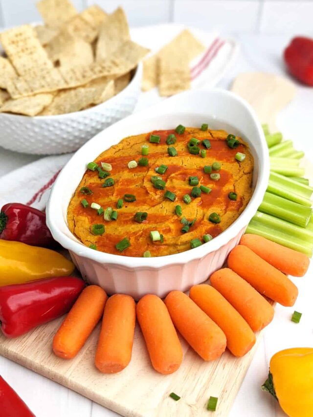 Buffalo chickpea dip in a casserole dish, surrounded by tortilla chips, carrot sticks, celery sticks, and fresh peppers.