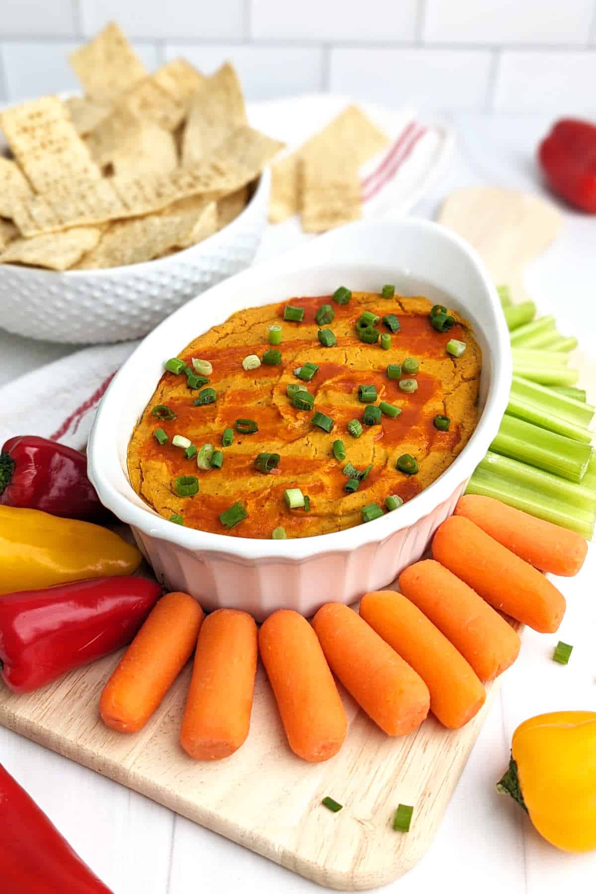 Buffalo chickpea dip in a casserole dish, surrounded by tortilla chips, carrot sticks, celery sticks, and fresh peppers.
