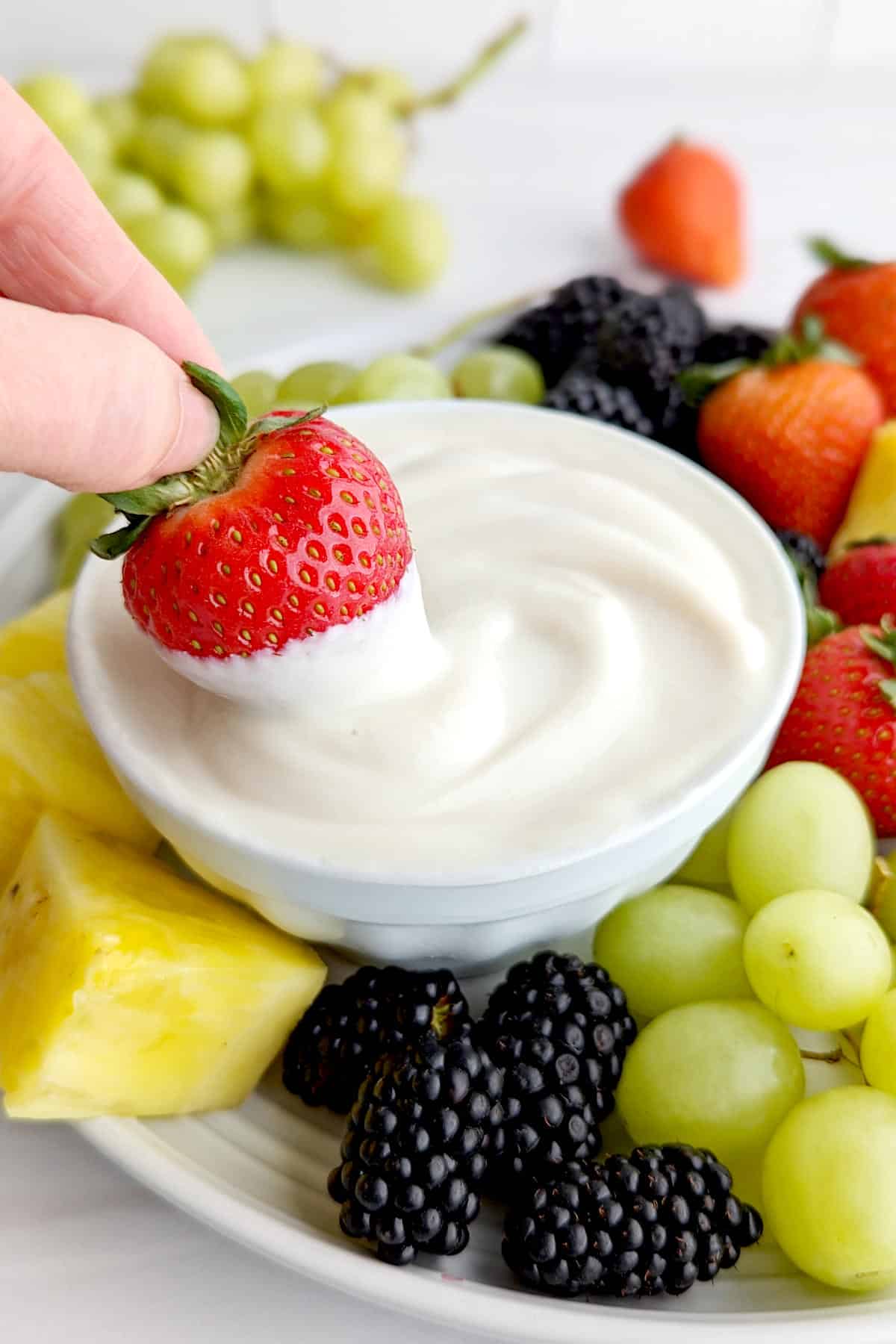 A hand dipping a strawberry into a bowl of vegan fruit dip.