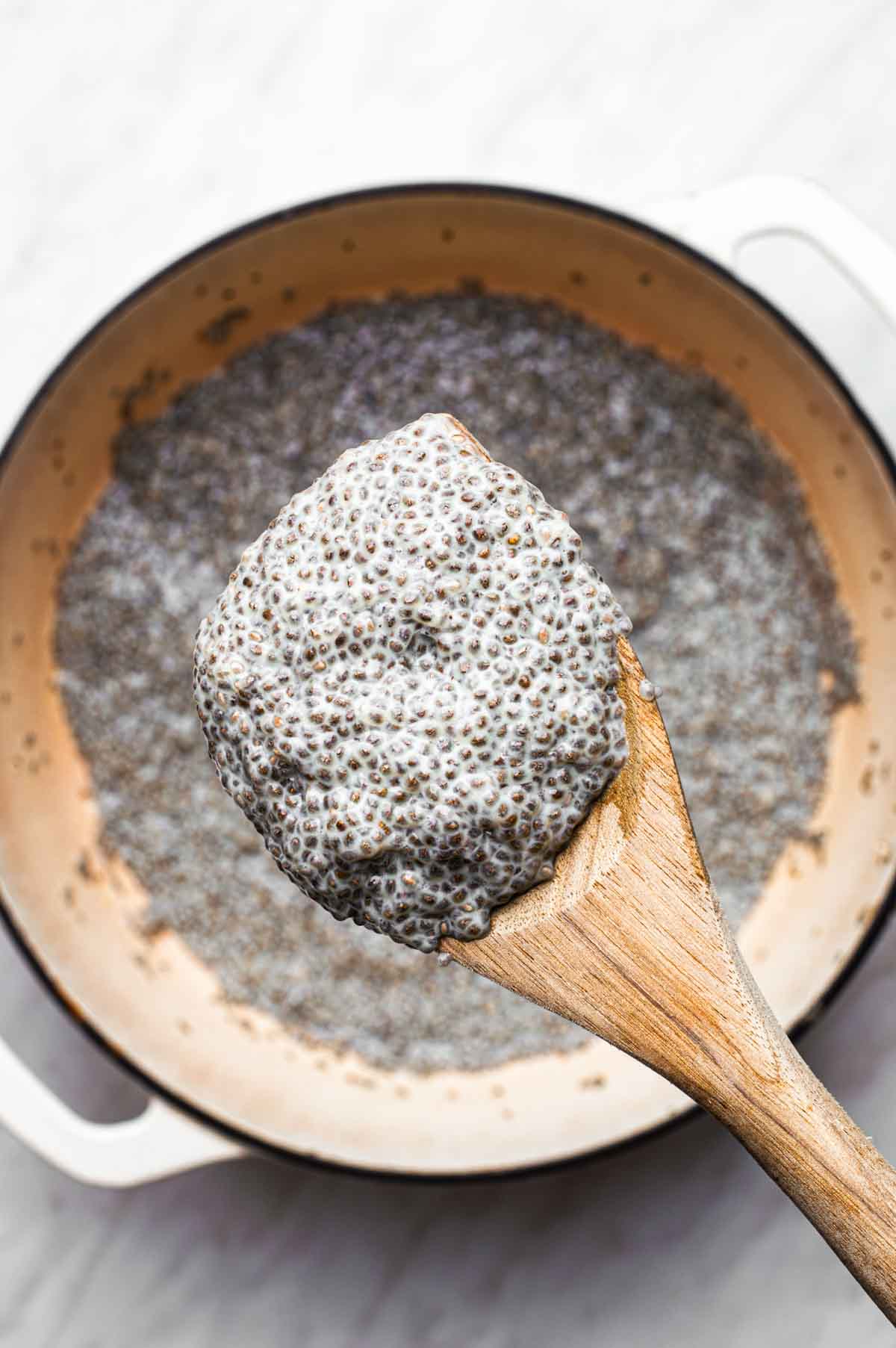 Thickened warm chia pudding on a cooking spoon hovering over a saucepan of it.