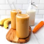 Carrot banana smoothie into two glasses with glass straws, next to ripe bananas and a raw carrot.