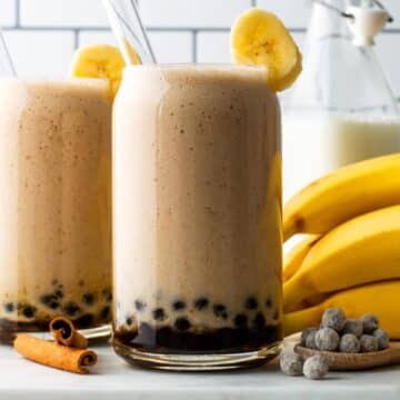 Two glasses filled with banana boba tea and served with banana slices on the rims and glass straws.
