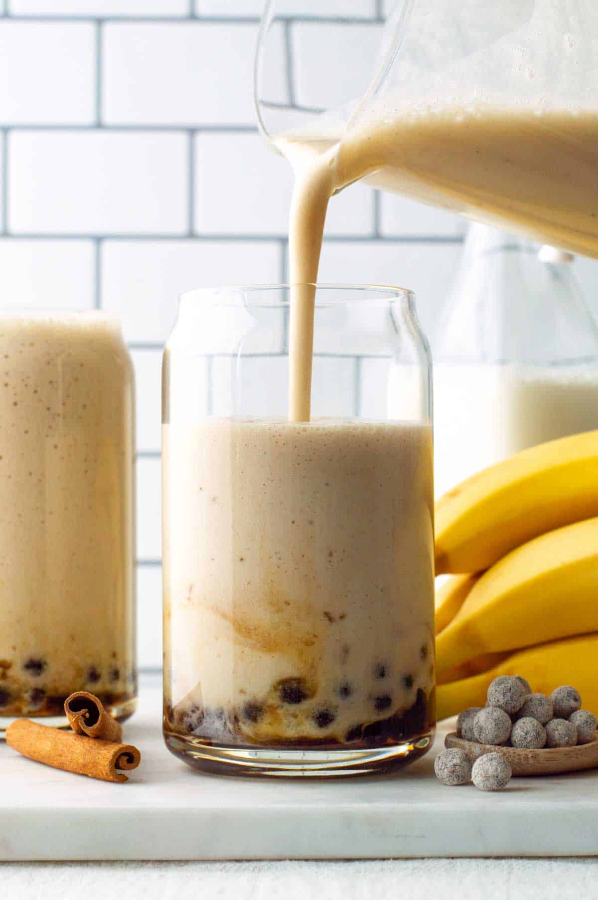 A pitcher pouring banana milk over boba pearls in a glass.