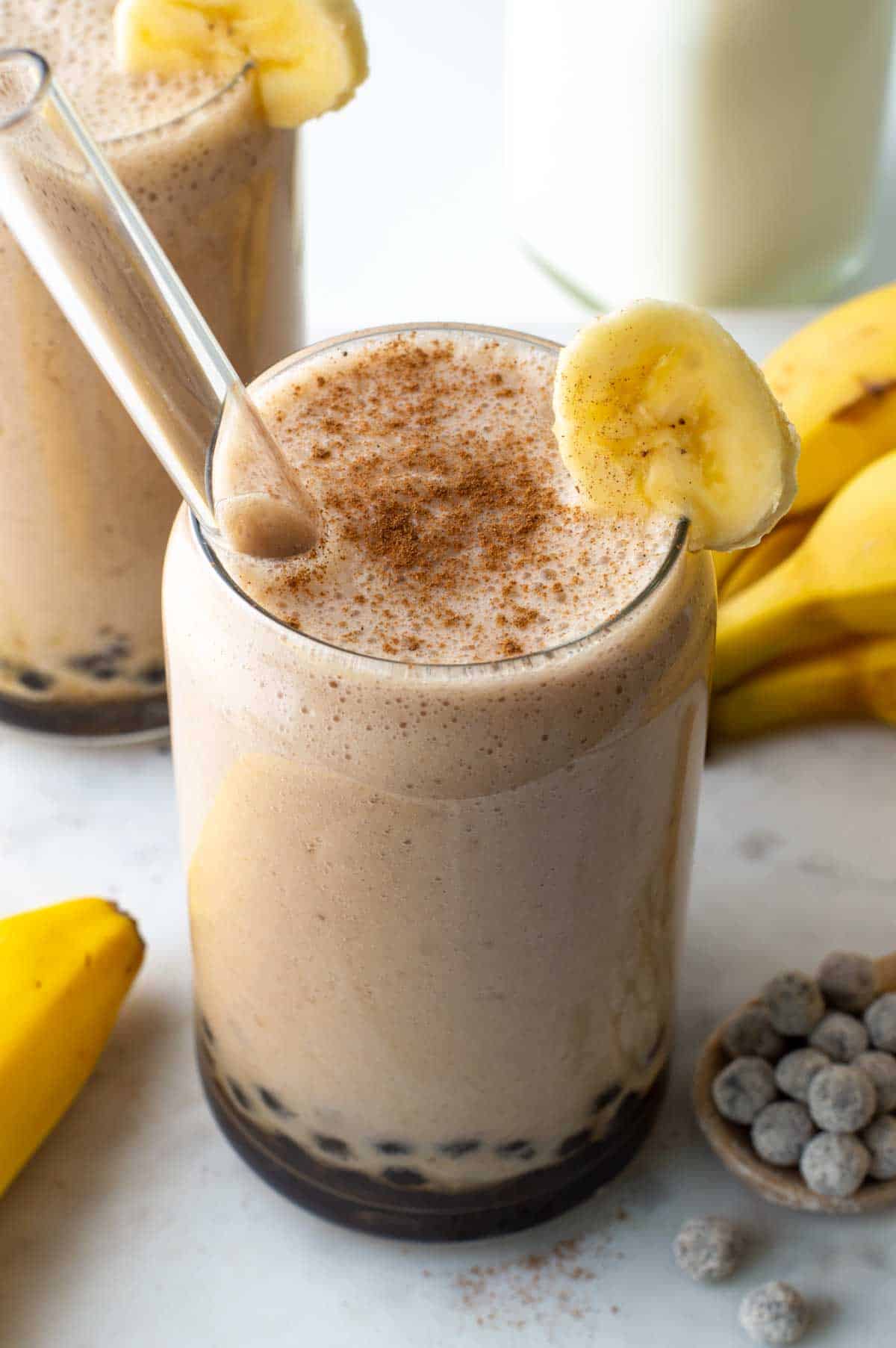 Banana milk boba tea served in a glass with a sprinkle of cinnamon on top and a banana slice on the rim.