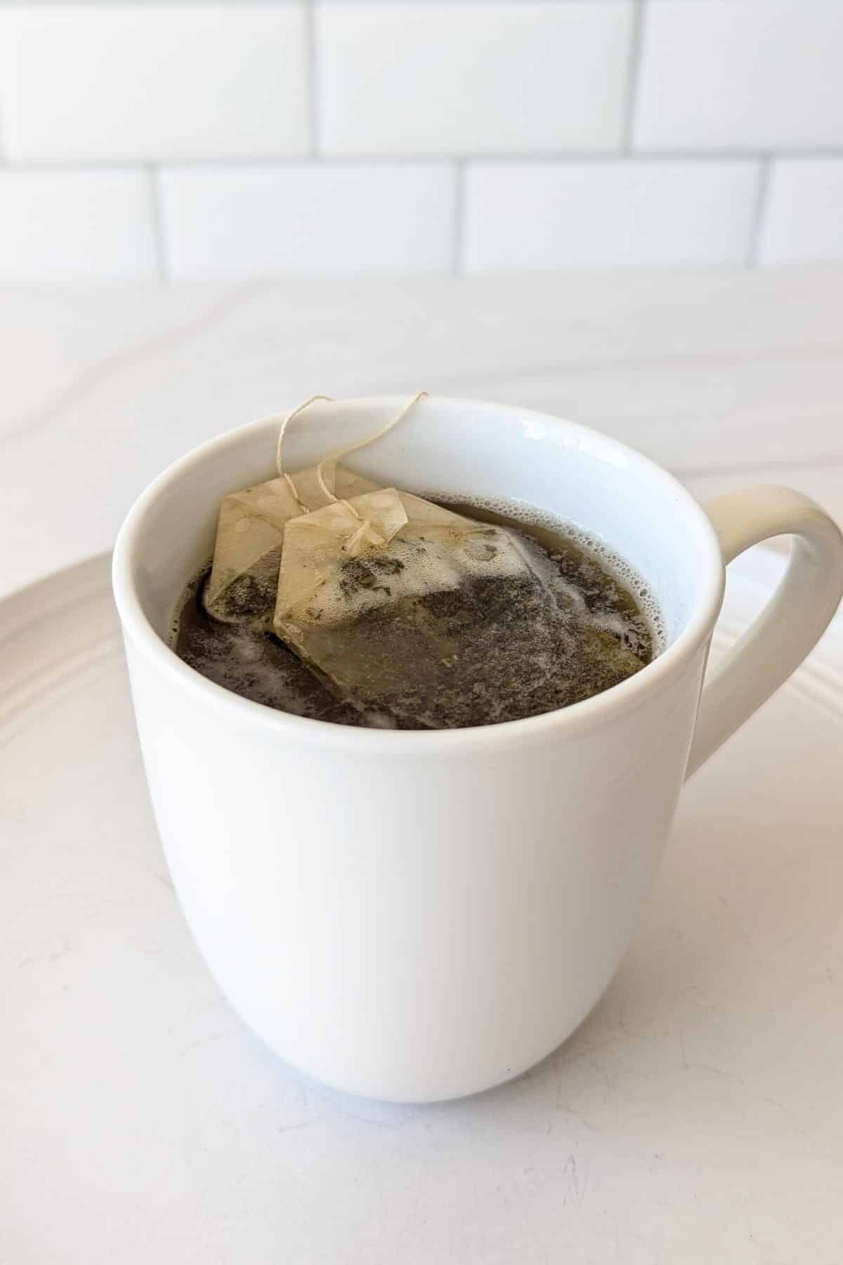 Two black tea bags steeping in hot water in a white mug sitting on a plate.