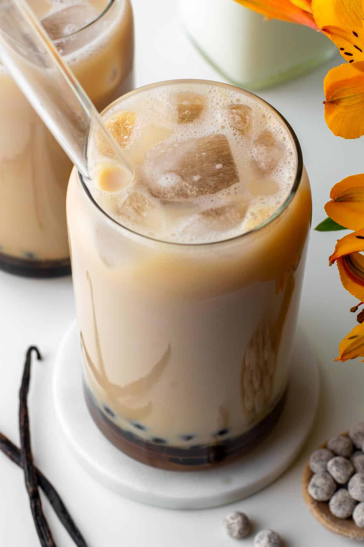 A glass of vanilla milk tea with boba pearls in the bottom, served with ice and a glass straw.
