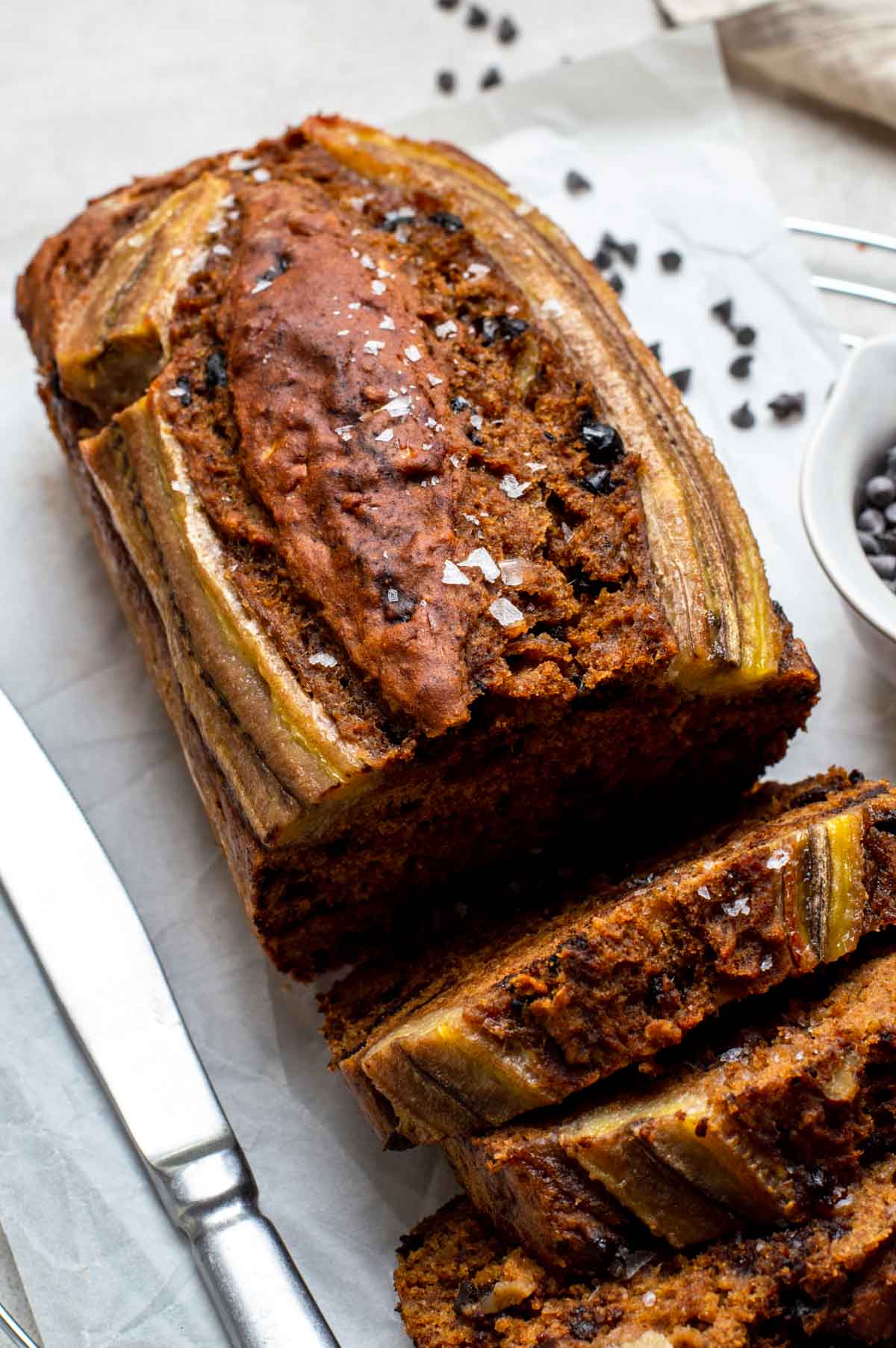 A partially sliced loaf of healthy vegan banana bread topped with flaky sea salt, sitting next to a bowl of chocolate chips.