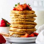 A tall stack of vegan bisquick pancakes on a plate, topped with fresh strawberries and maple syrup.