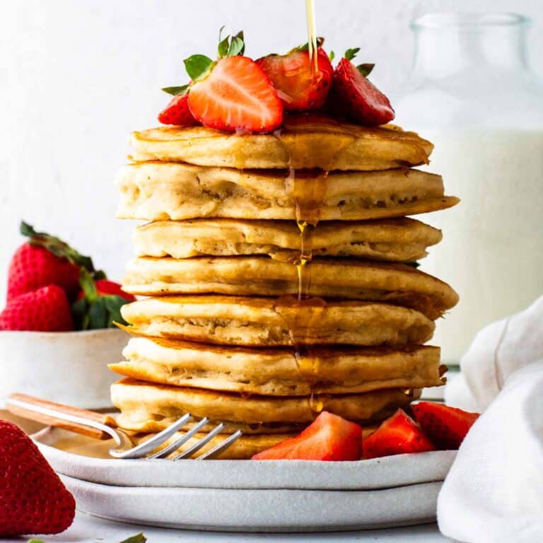 A tall stack of vegan bisquick pancakes on a plate, topped with fresh strawberries and maple syrup.