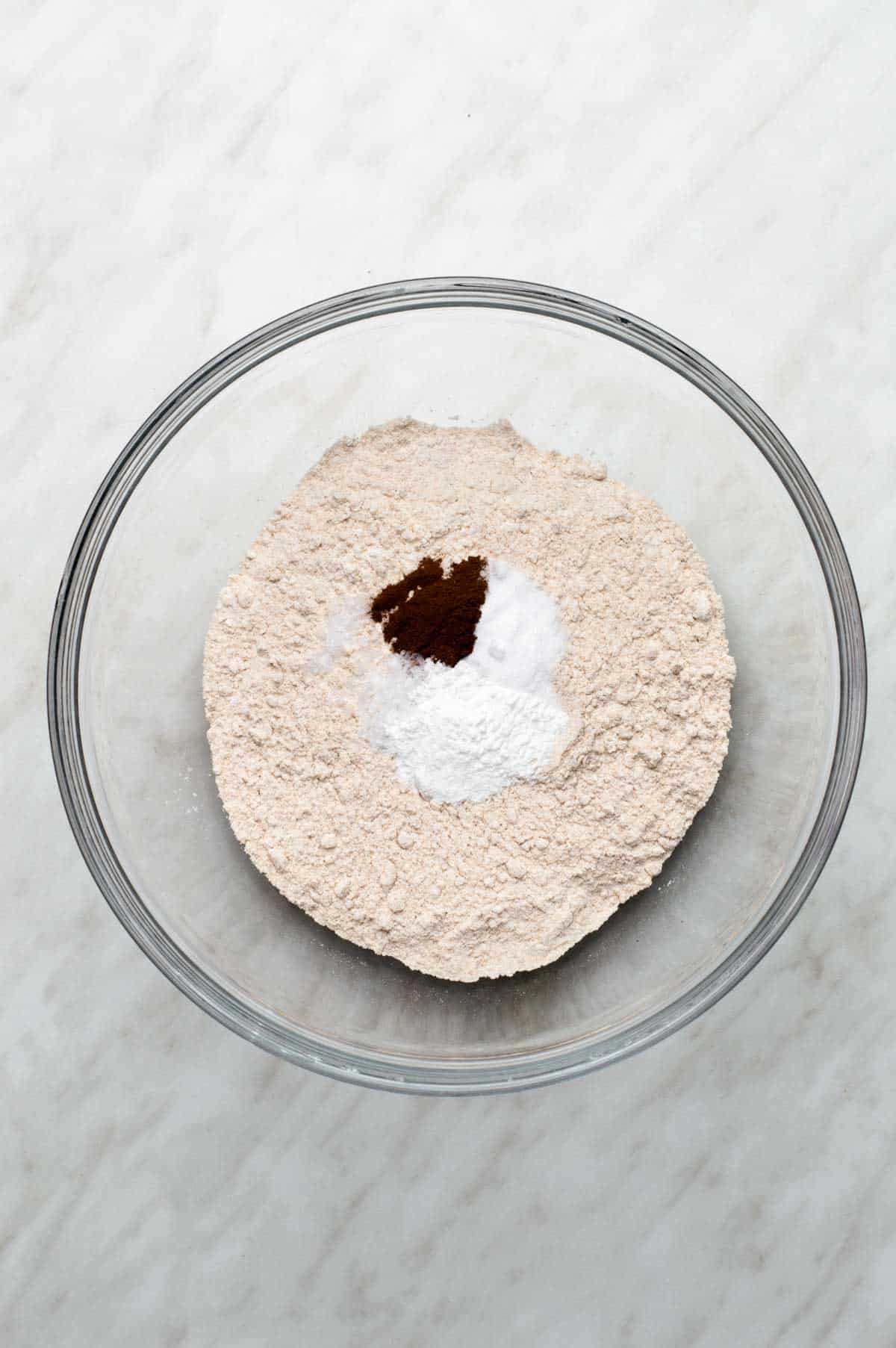A clear glass bowl filled with flour, baking powder, baking soda, salt, and cinnamon, arranged in a small mound before mixing.
