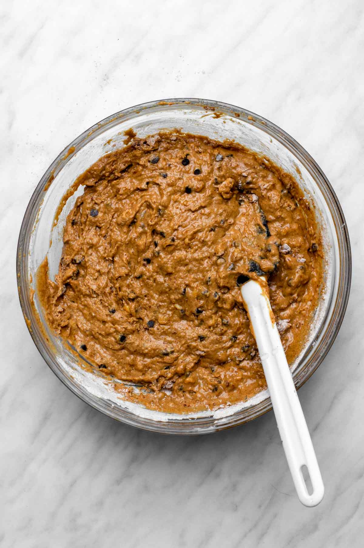 A glass mixing bowl filled with thick banana bread batter, speckled with chocolate chips, and a white spatula resting on the edge.
