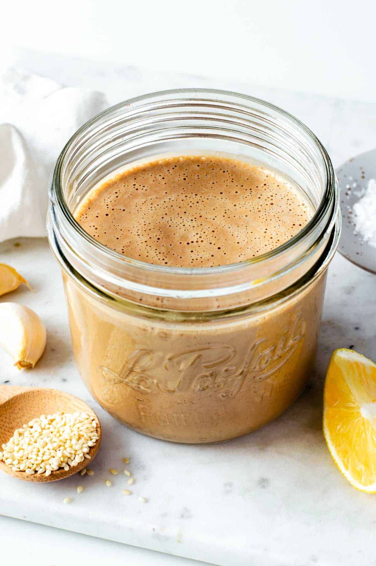 A small glass jar filled with creamy tahini balsamic dressing, next to sesame seeds and a lemon wedge.