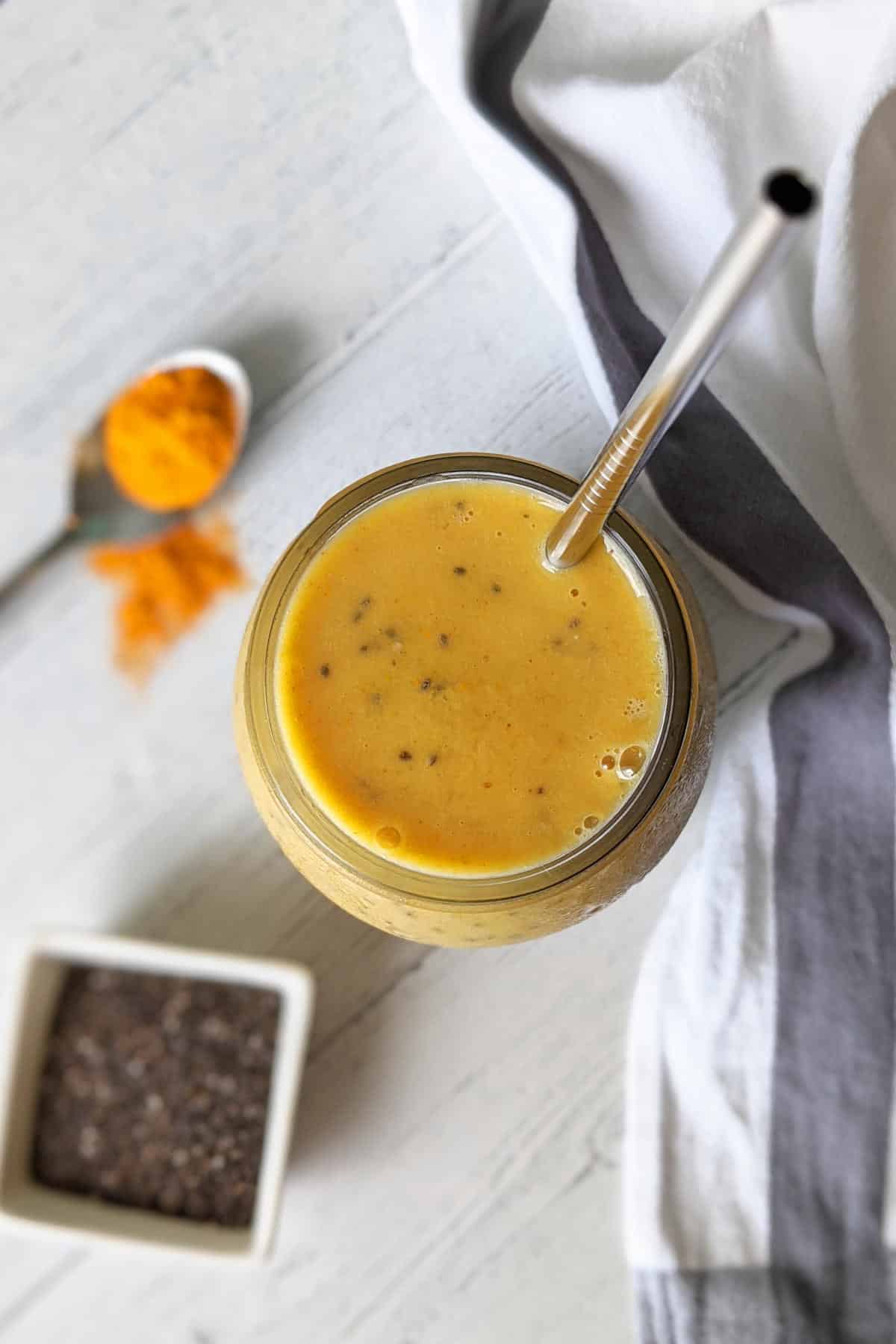 Maca smoothie made with bananas and mangos in a glass with a straw, next to a spoon of turmeric powder and a bowl of chia seeds.