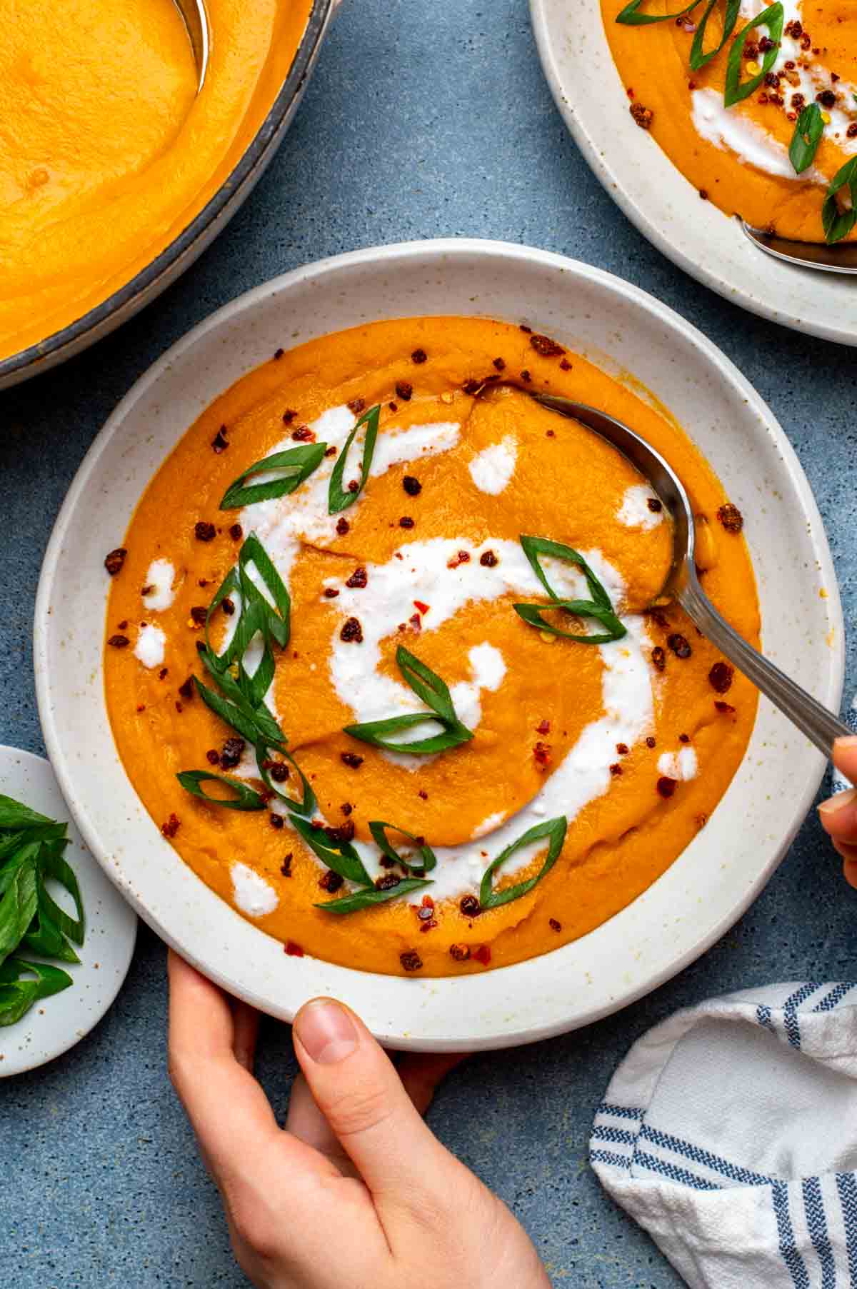 A hand reaching towards a bowl of carrot and lentil soup topped with a swirl of coconut milk and sliced green onions.