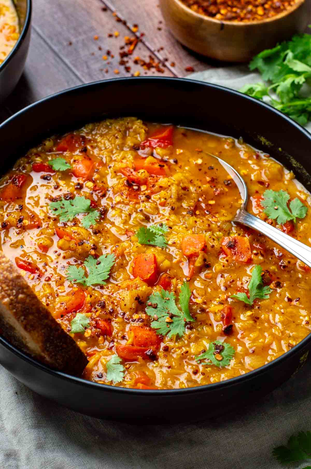 Bowl of lentil red pepper soup with a spoon inside, served with a slice of bread on the side.