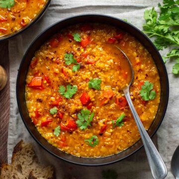 Bowl of lentil red pepper soup with a spoon resting in it, garnished with fresh herbs.