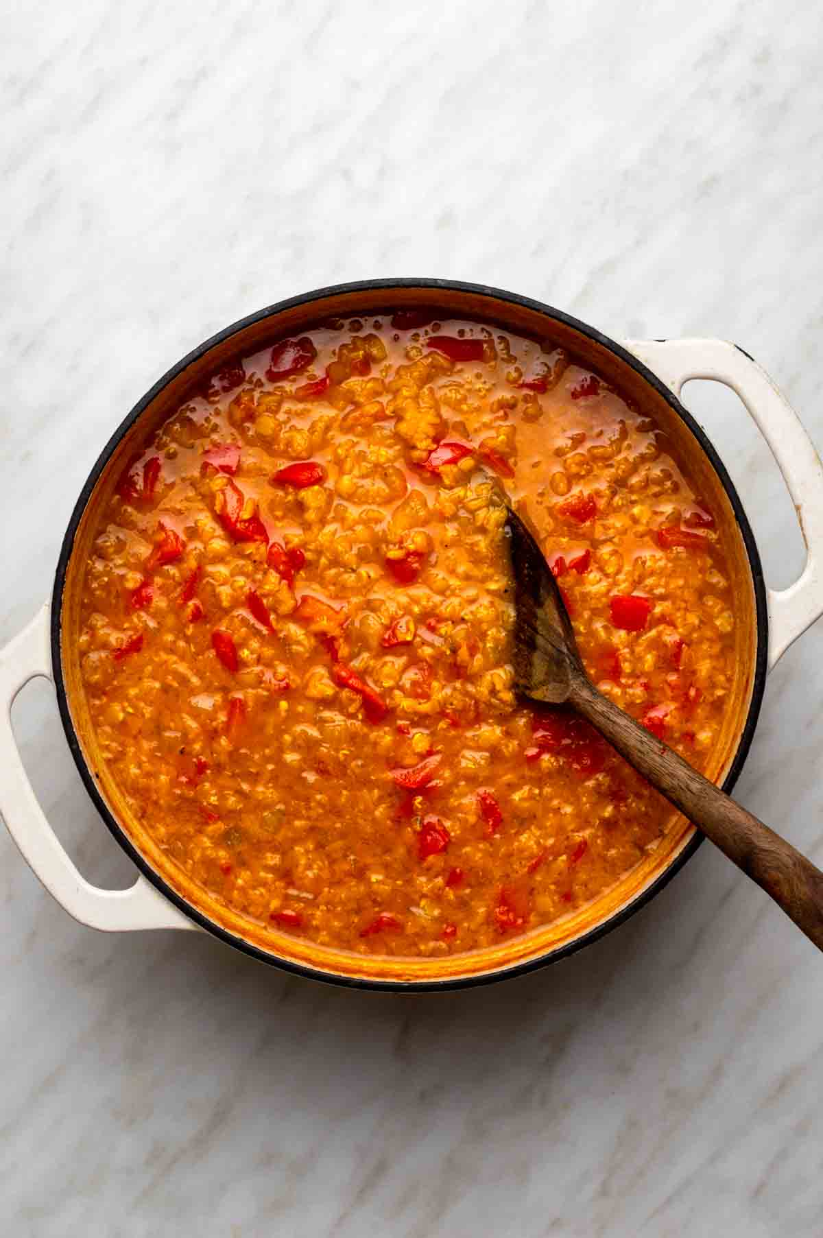 Large stockpot filled with diced bell peppers, onions, tomato paste, spices, vegetable broth, and red lentils being stirred together.
