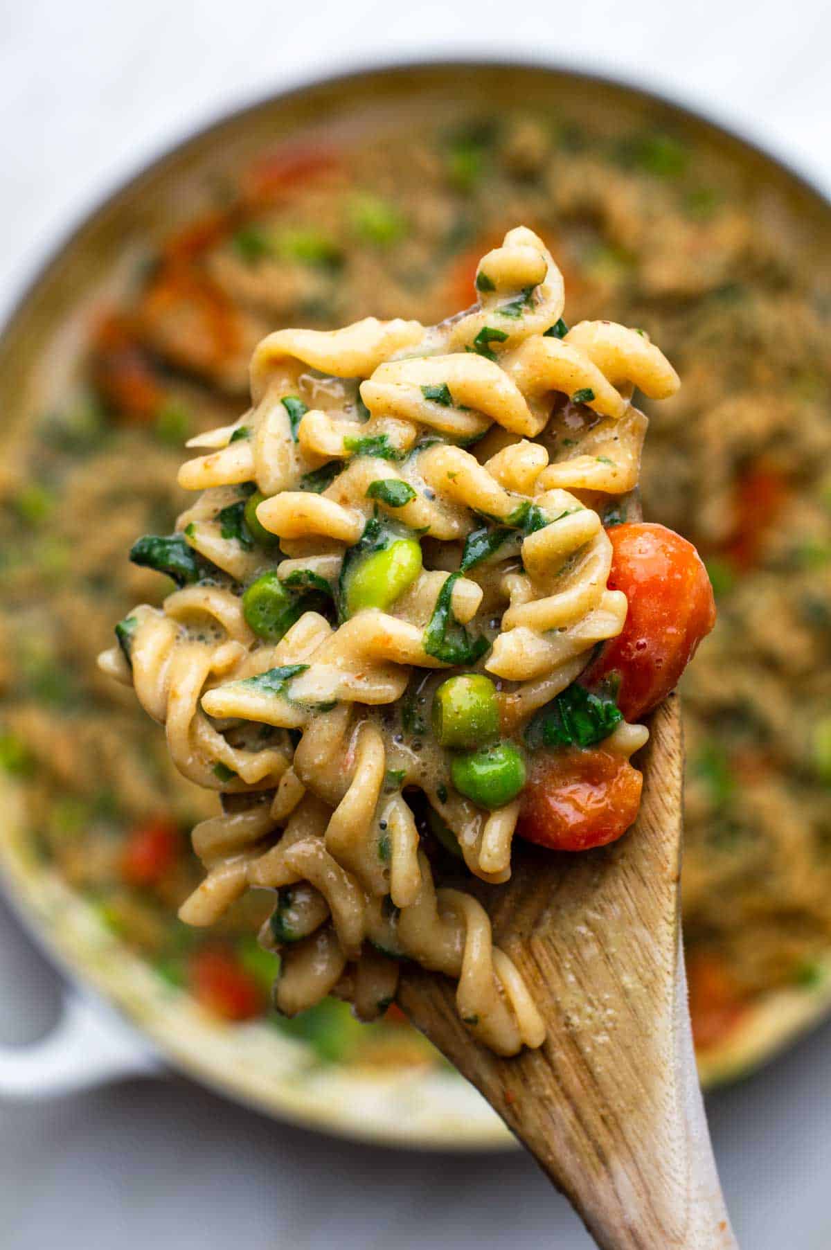A wooden spoon lifting a scoop of creamy veggie pasta from the pan.