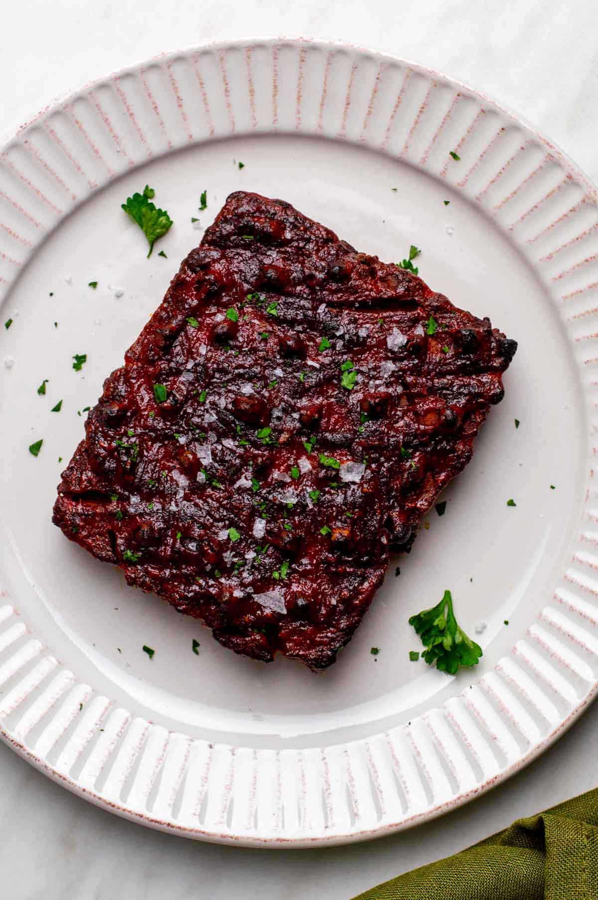 A single square piece of finished vegan ham with a glossy, dark red glaze, garnished with salt flakes and parsley on a white plate.