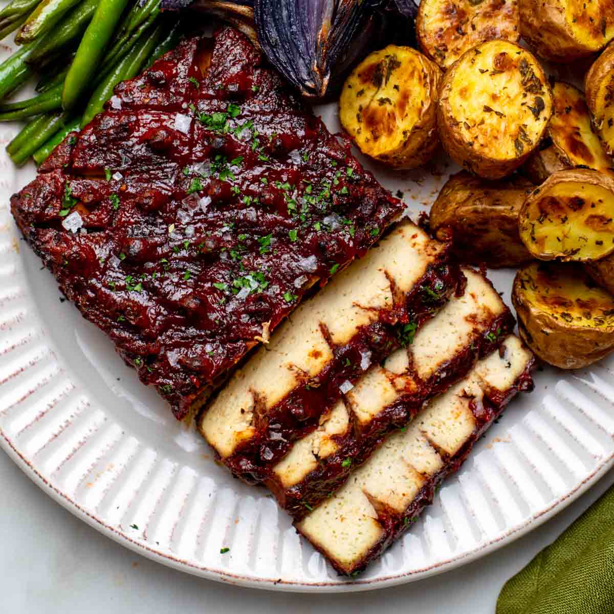 Sliced vegan ham with a red glaze, served with roasted potatoes, green beans, and charred red onions.