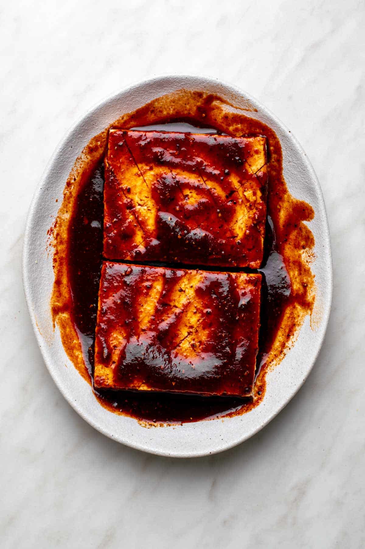Two scored blocks of tofu saturated in a thick, deep red marinade on a white plate.