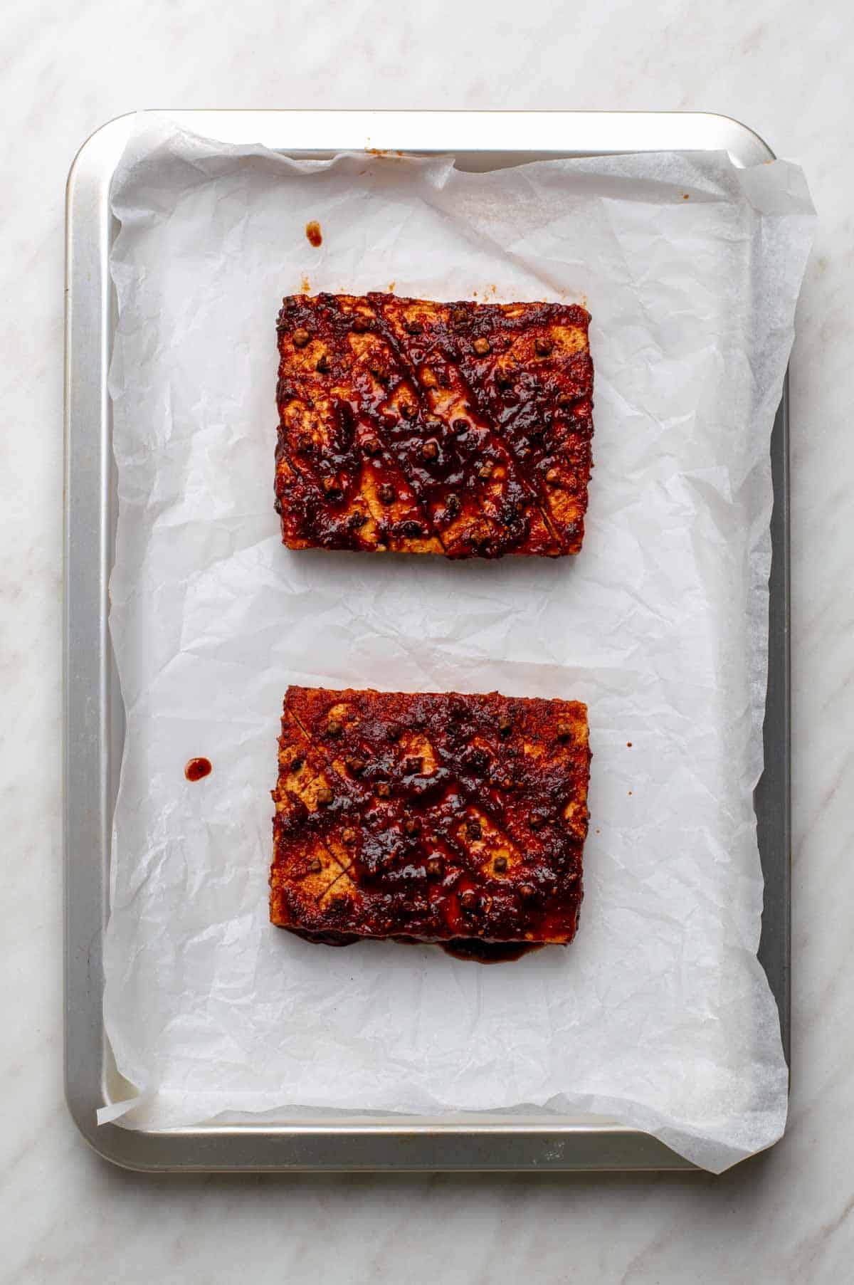 Two marinated and scored tofu blocks placed on a parchment-lined baking sheet ready for baking.