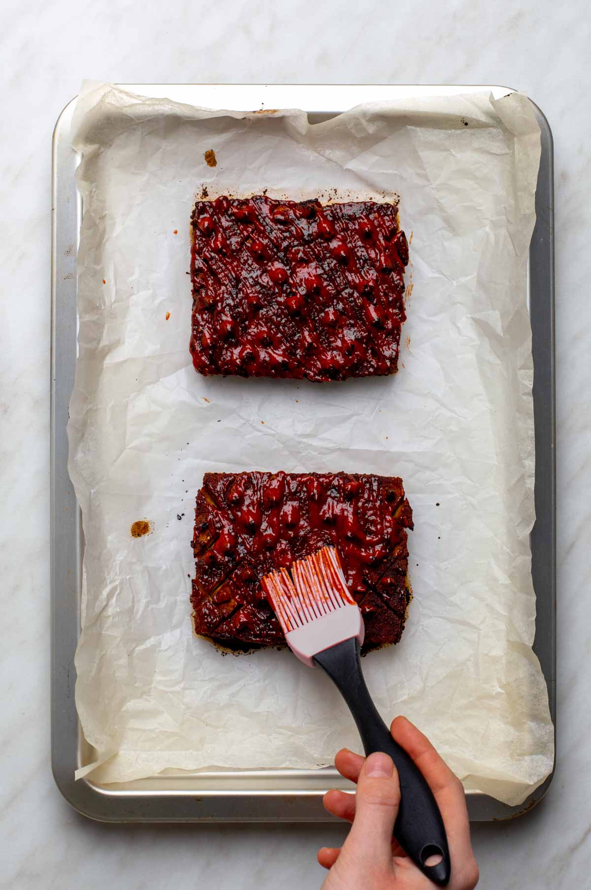 A hand brushing a red, sticky glaze onto two scored squares of vegan ham on a parchment-lined baking sheet.