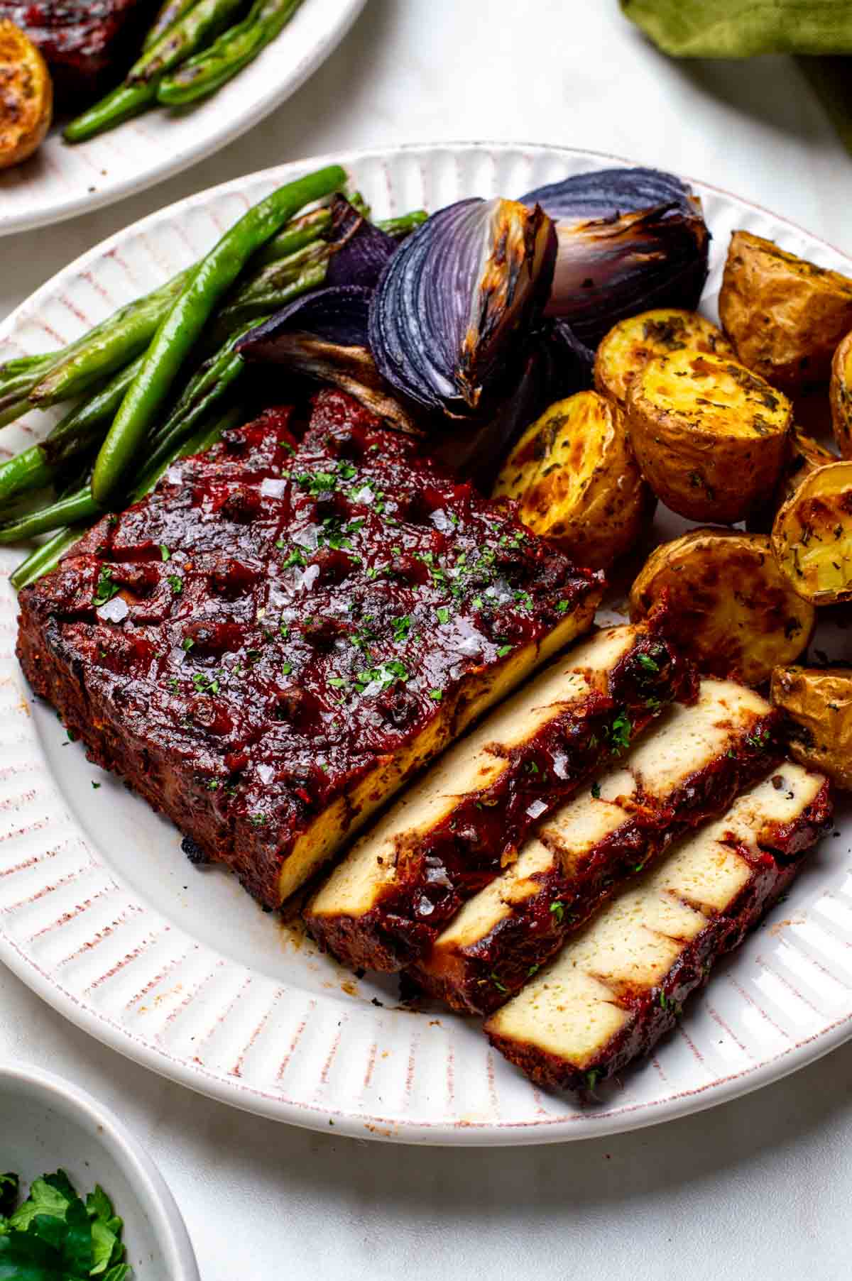 Sliced vegan ham with a red glaze, served with roasted potatoes, green beans, and charred red onions.