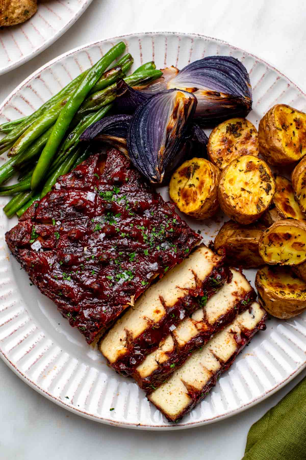 Sliced vegan ham with a red glaze, served with roasted potatoes, green beans, and charred red onions.