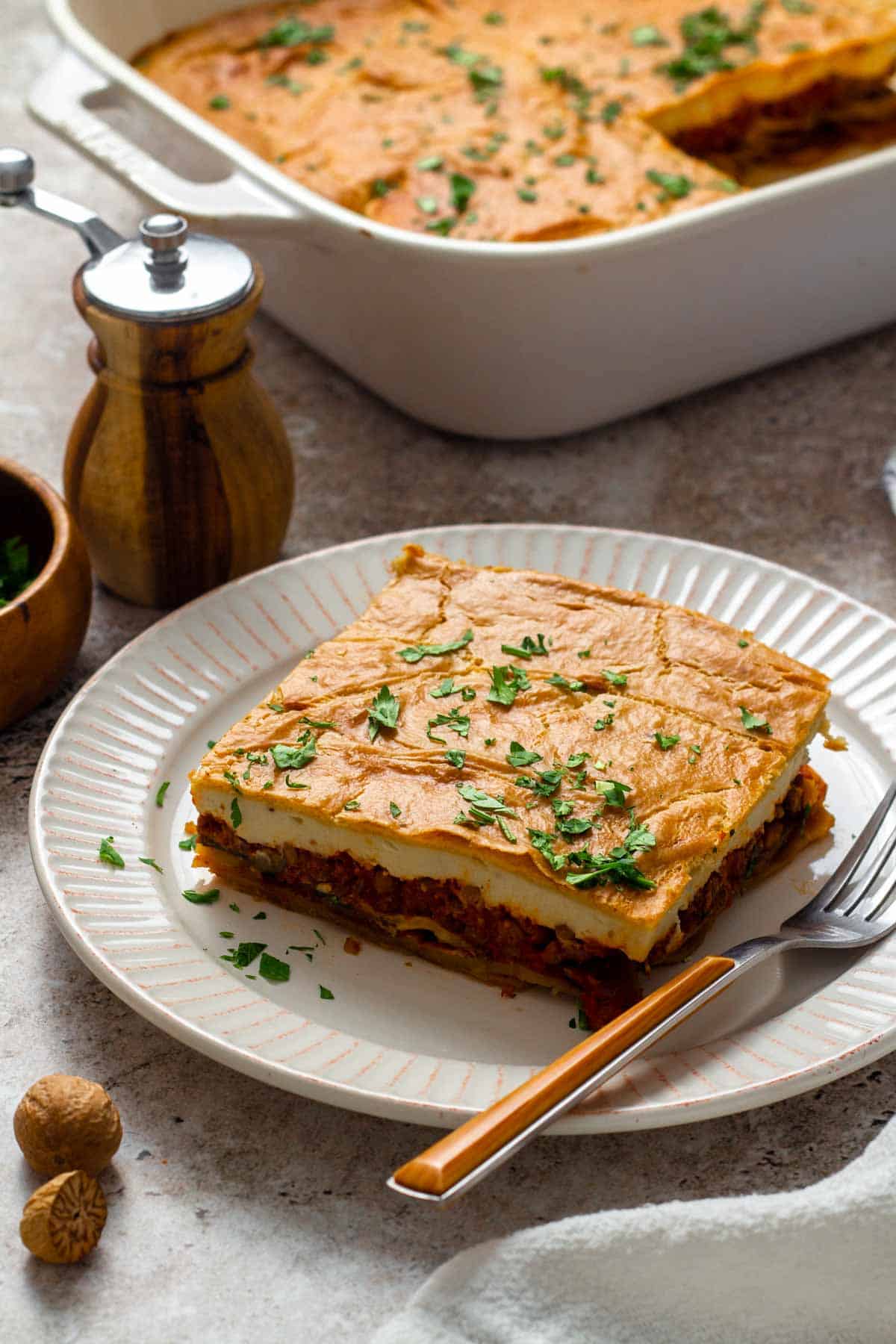 A slice of vegan moussaka on a white plate, showing layers of roasted eggplant, tomato-lentil filling, and creamy cashew b&eacute;chamel, garnished with chopped parsley.