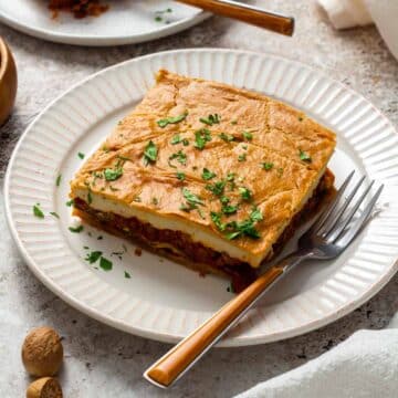 A slice of vegan moussaka on a white plate, showing layers of roasted eggplant, tomato-lentil filling, and creamy cashew béchamel, garnished with chopped parsley.