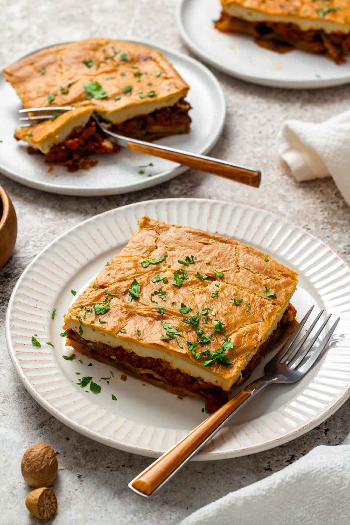 A slice of vegan moussaka on a white plate, showing layers of roasted eggplant, tomato-lentil filling, and creamy cashew béchamel, garnished with chopped parsley.